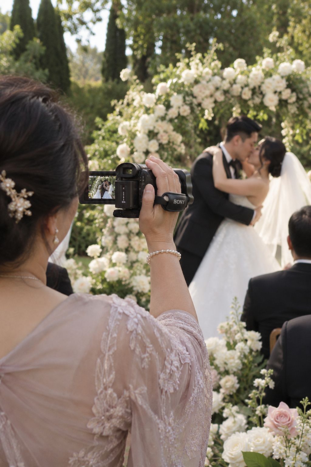 A wedding guest in a lace dress films the couple's first kiss on a handheld camcorder